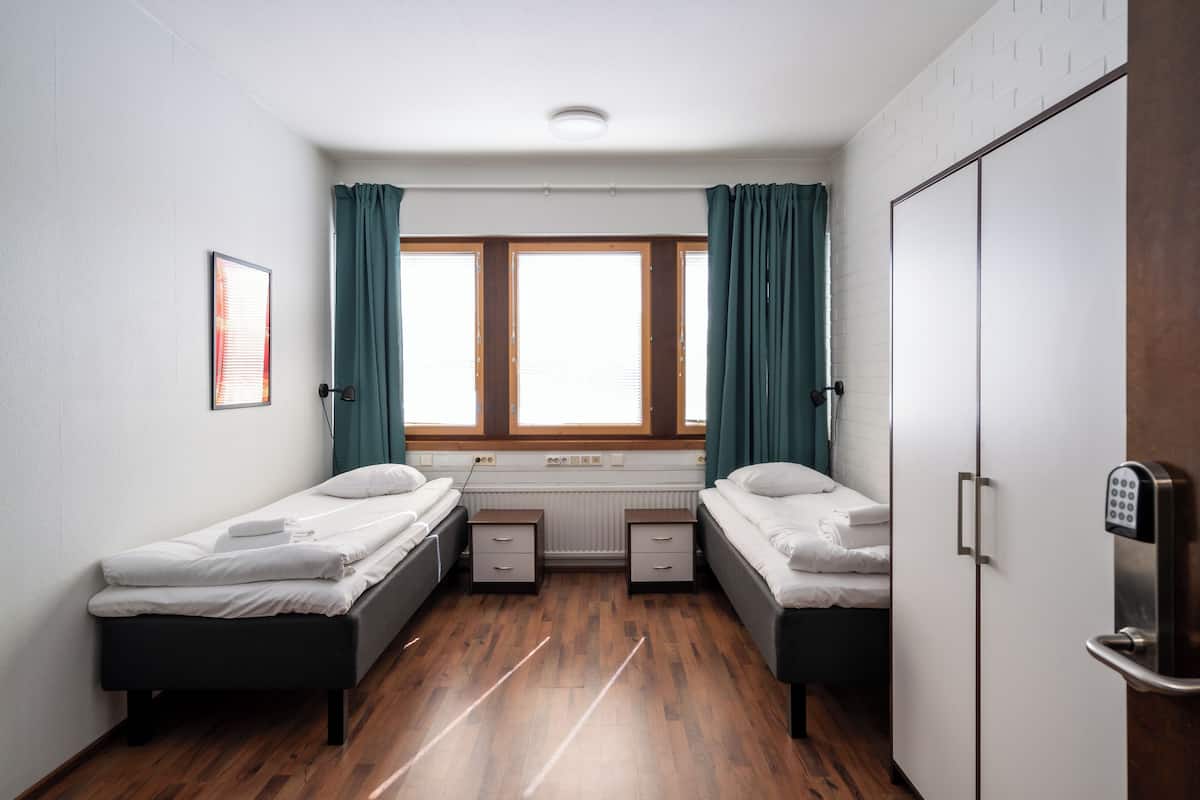A small, clean room with two single beds, a window with curtains, a radiator, and a wardrobe.