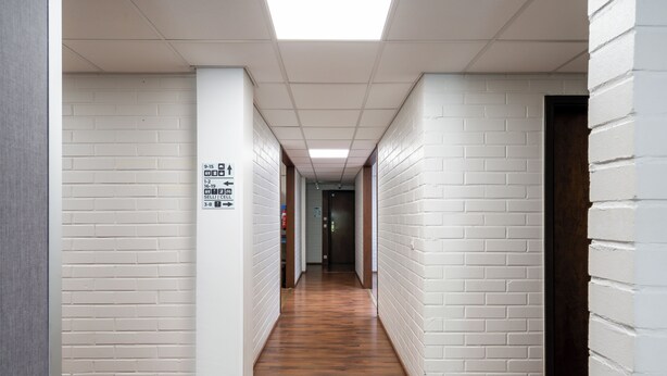 A hallway with white brick walls, wooden flooring, and a ceiling with fluorescent lights. - Hiisi Hostel Vihti - Private Rooms (Vihti)
