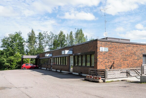 A brick building with a covered parking area and a red car parked outside. - Hiisi Hostel Vihti - Private Rooms (Vihti)