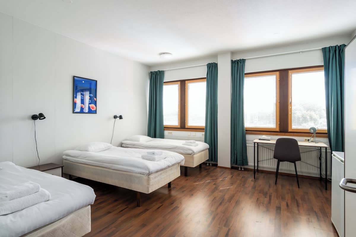 A hotel room with two single beds, a desk with a chair, a radiator, and a window with curtains.