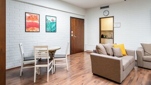 A modern living room with a white brick wall, a wooden floor, a round table with chairs, a beige sofa with cushions, and a clock on the wall. - Hiisi Hostel Vihti - Private Rooms (Vihti)
