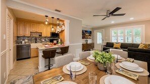 A modern kitchen with wooden cabinets, a dining area with a round table set for four, and a living room with a dark sofa and a ceiling fan. - **SUNFLOWER MELODIES @ MIDTOWN** Home & Pool + Last Key White Glove Service... (Key West)