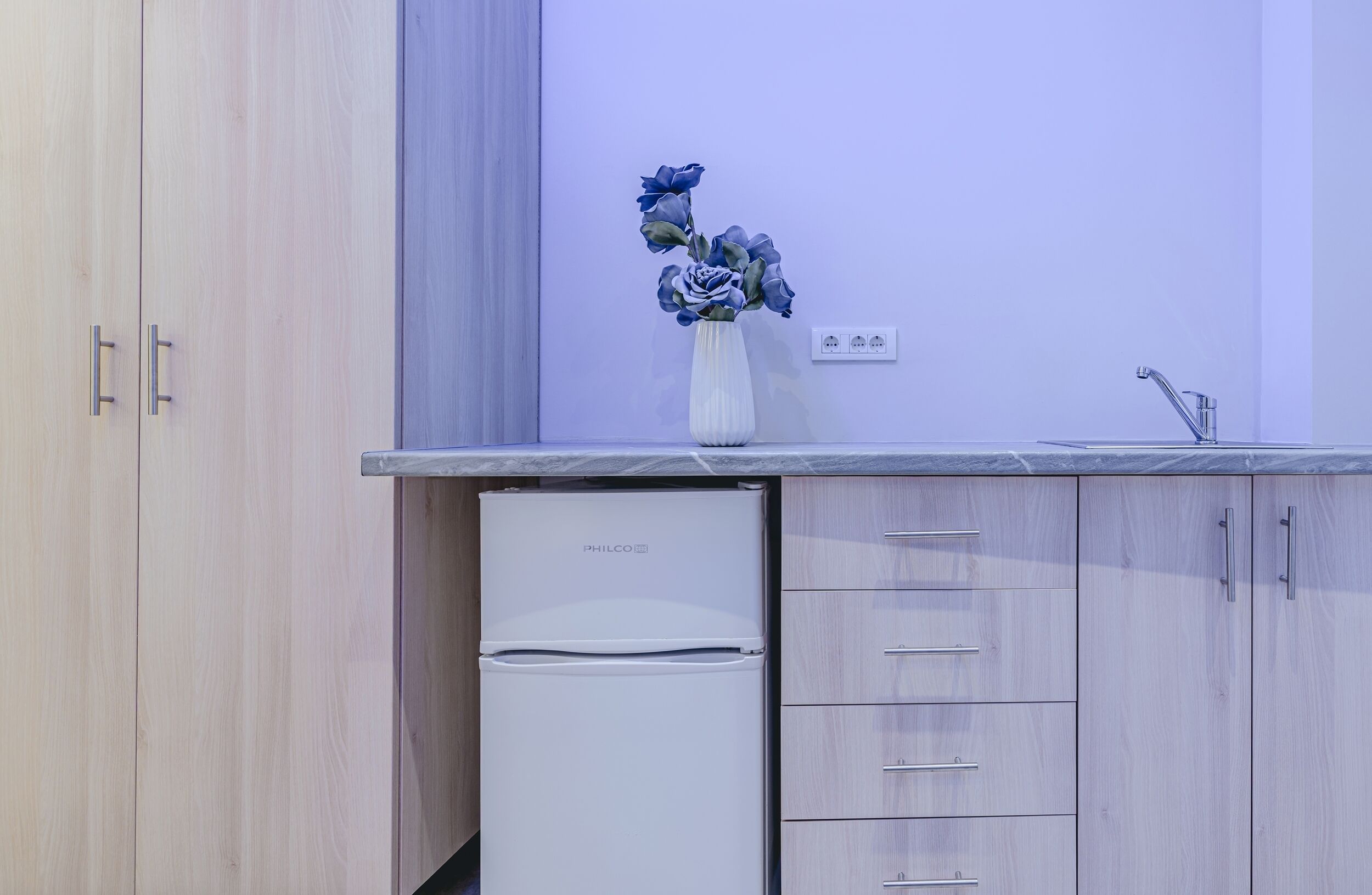 A compact kitchen with a PHILCO refrigerator, wooden cabinets, a white countertop, and a vase with flowers.