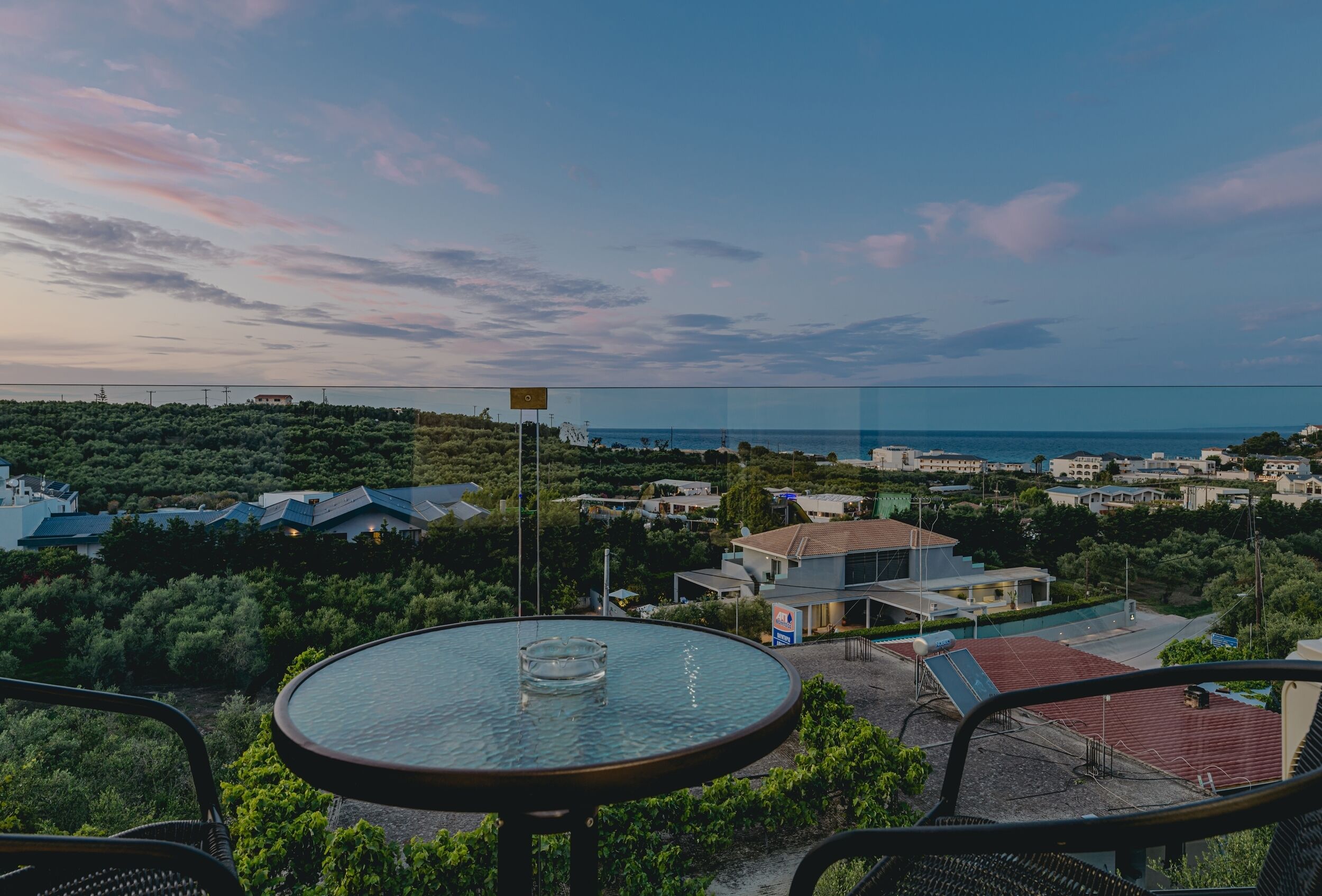 Eine Terrasse mit Glastisch, mit Blick auf eine Küstenlandschaft mit Gebäuden und Grünpflanzen.