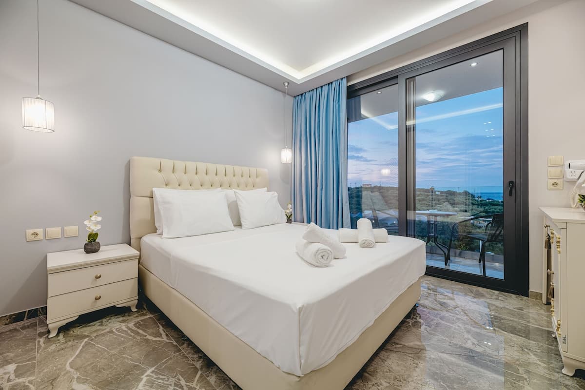 A modern hotel room with a large bed, a nightstand, a dresser, and a view of the city through a large window.