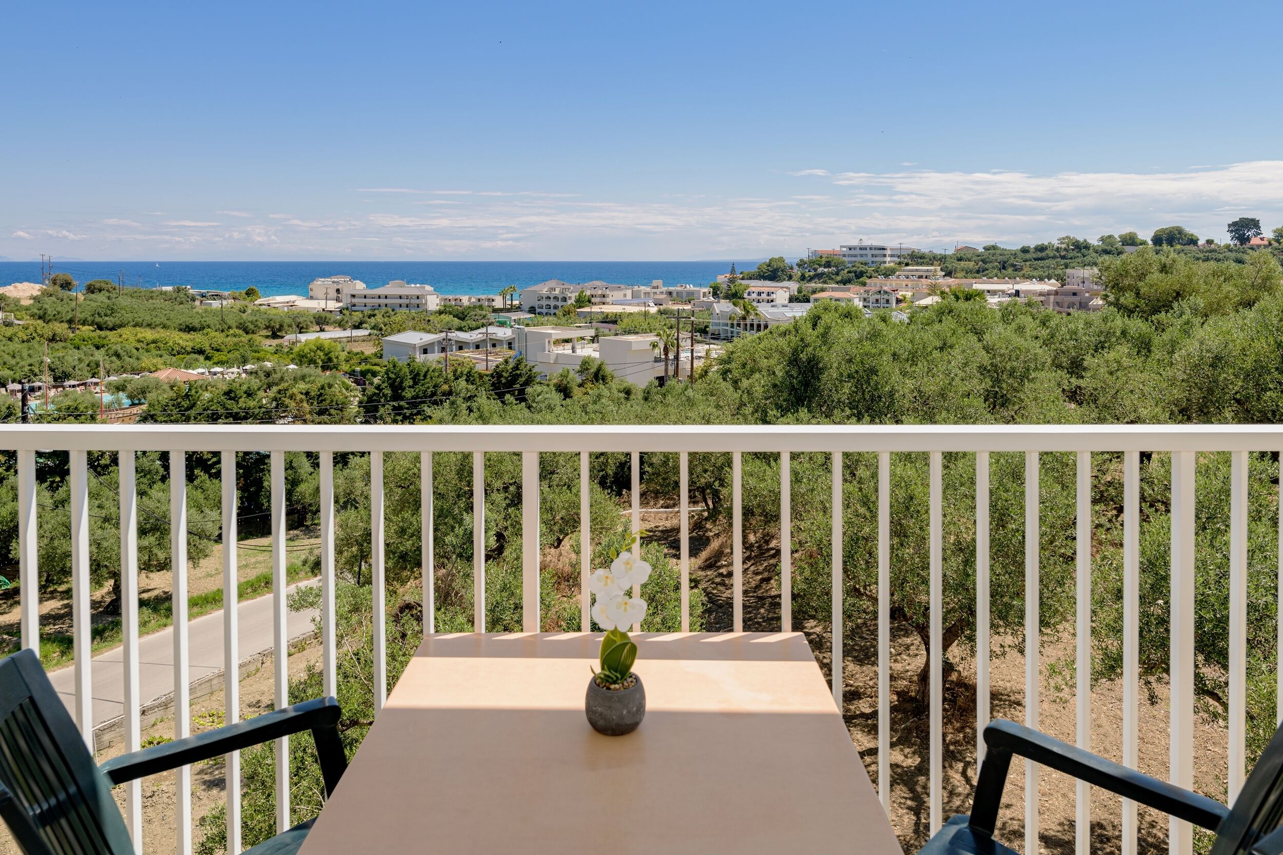 A balcony with a table and chairs, overlooking a coastal town with buildings and greenery.