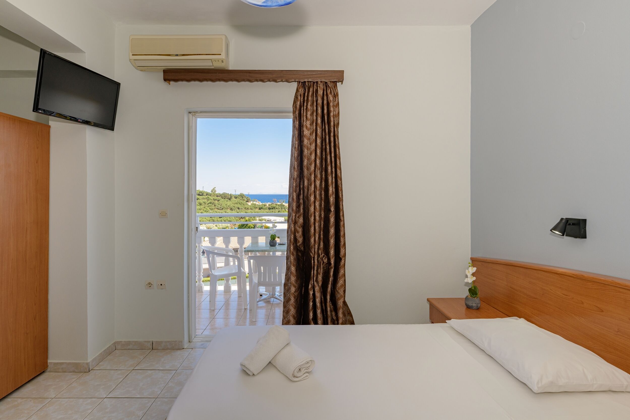 A hotel room with a bed, a TV, an air conditioner, a bedside table with a lamp, and a view of the outdoors.