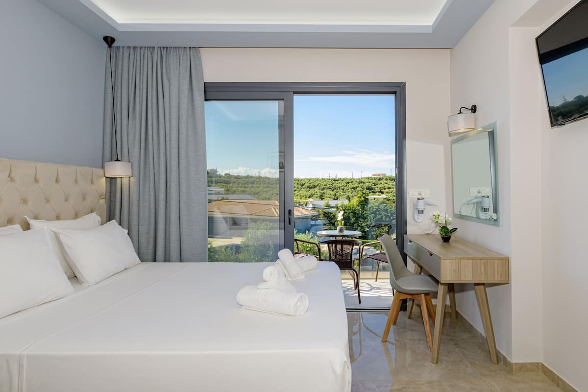 A hotel room with a large bed, a desk, a chair, a TV, and a balcony with a view of greenery.