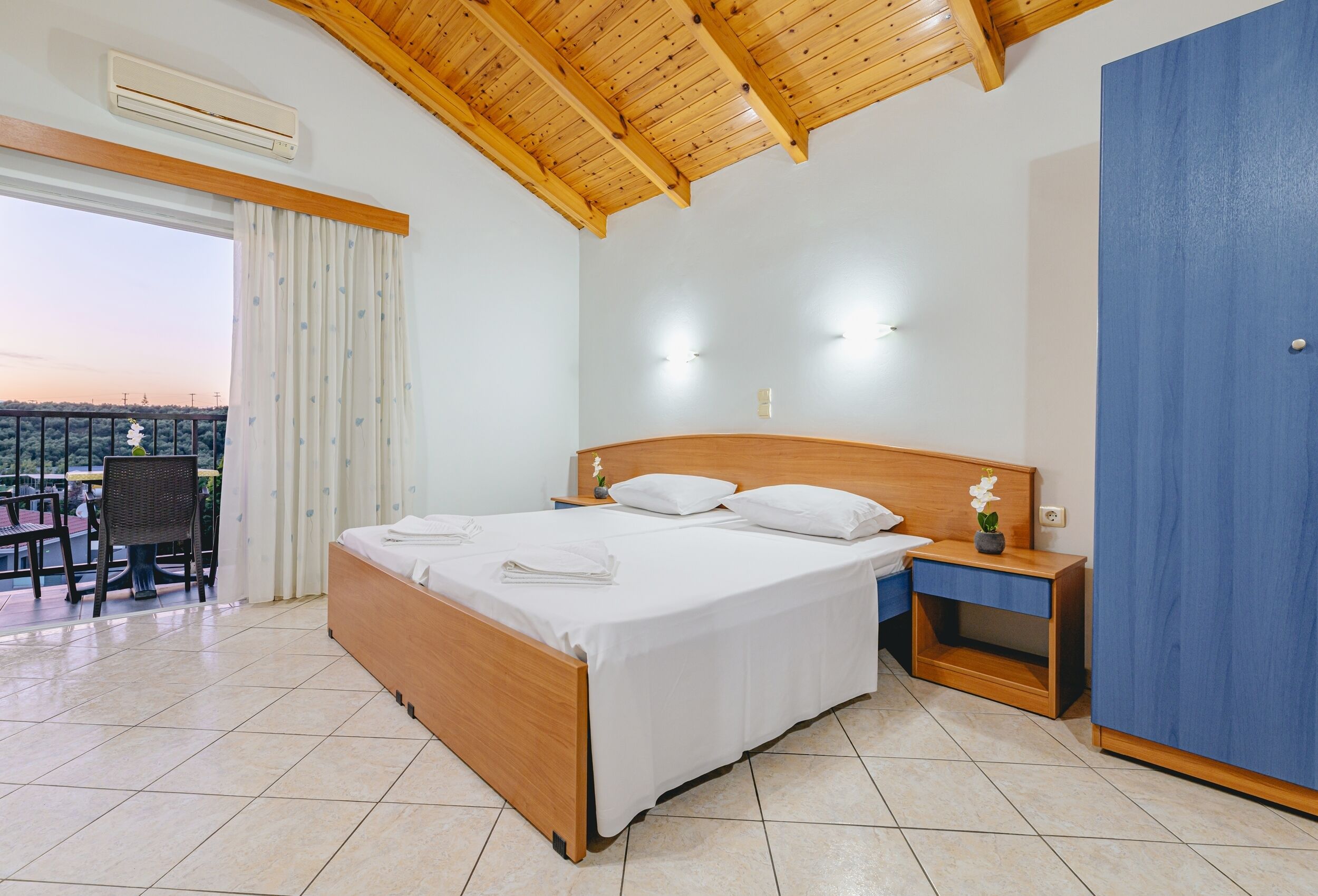 A bedroom with a wooden bed, a nightstand, a blue wardrobe, and a balcony with a chair.