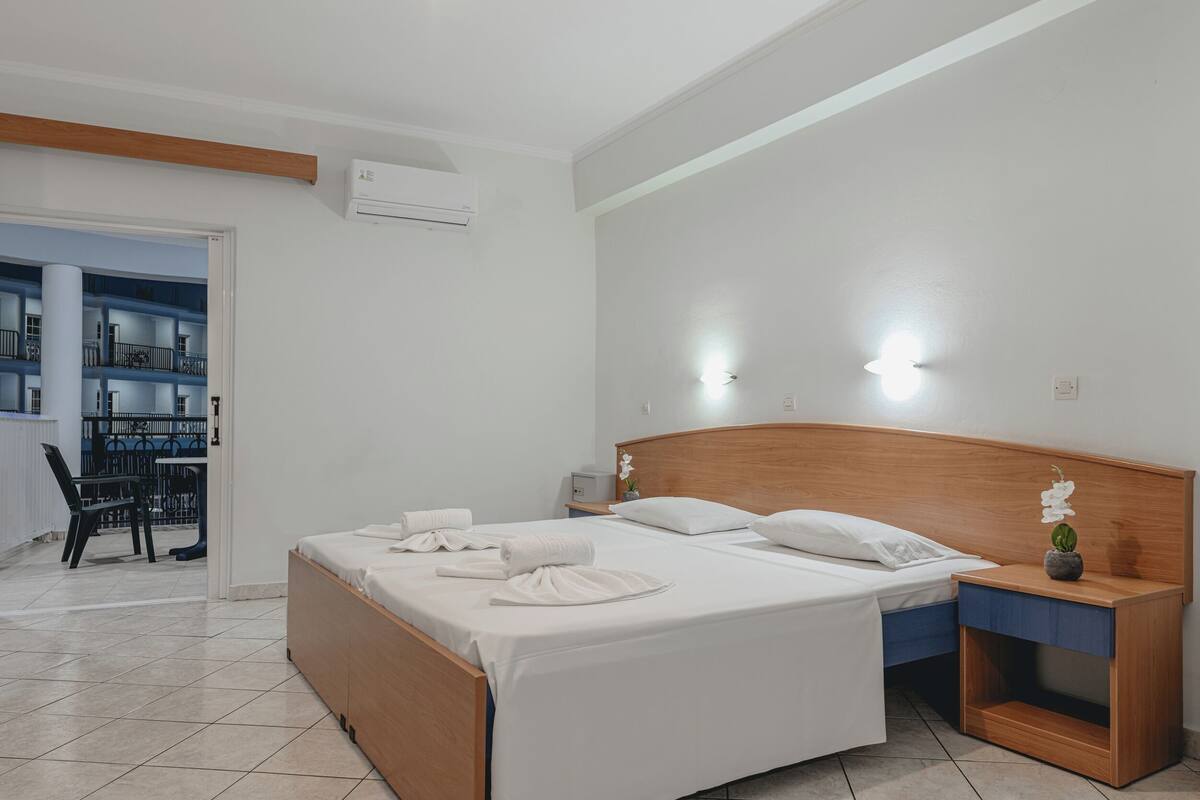 A modern hotel room with a wooden bed, white linens, a nightstand with a vase, and a view of a balcony with outdoor seating.