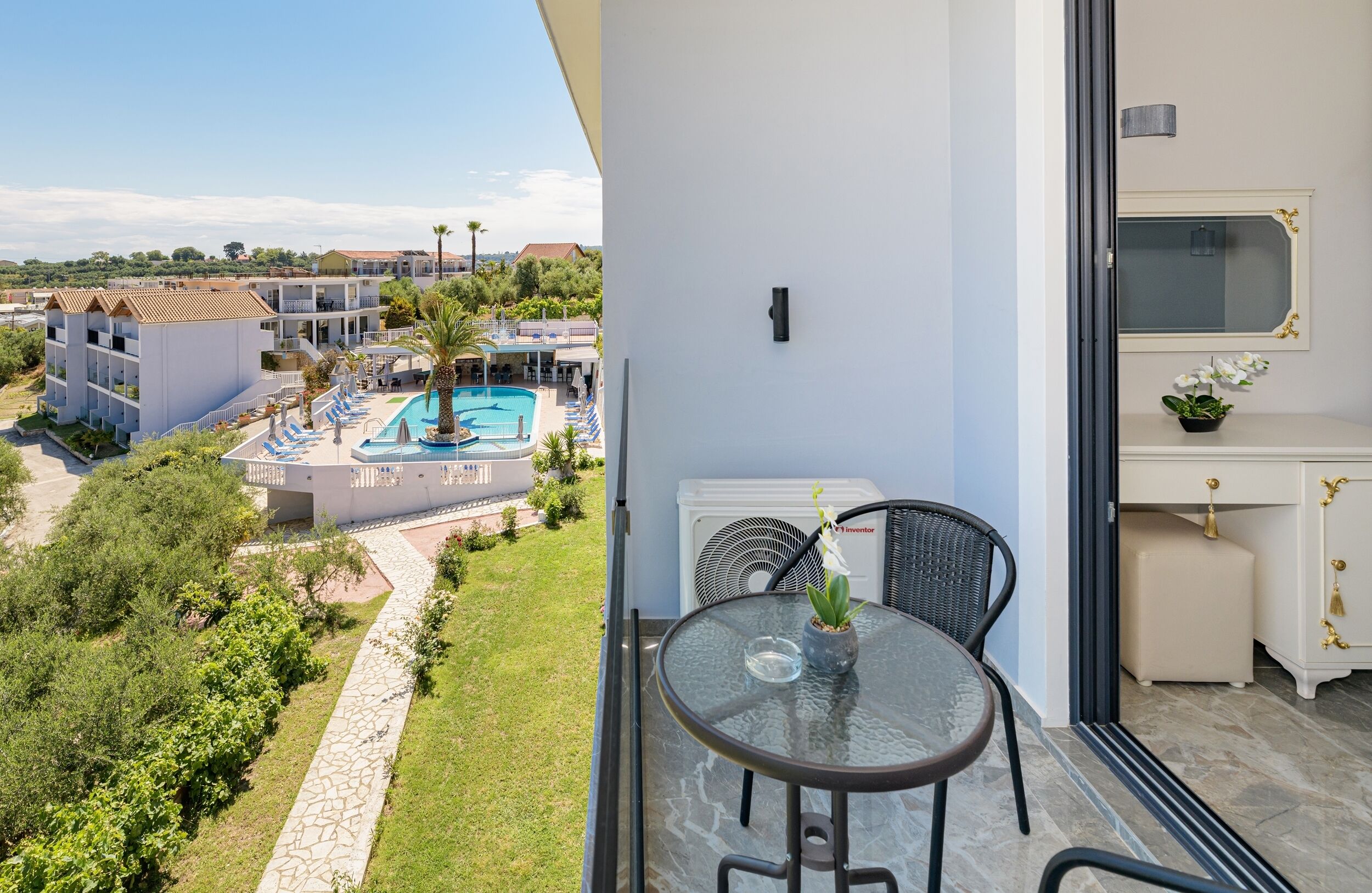 A balcony with a glass table and chairs, overlooking a swimming pool and hotel buildings.