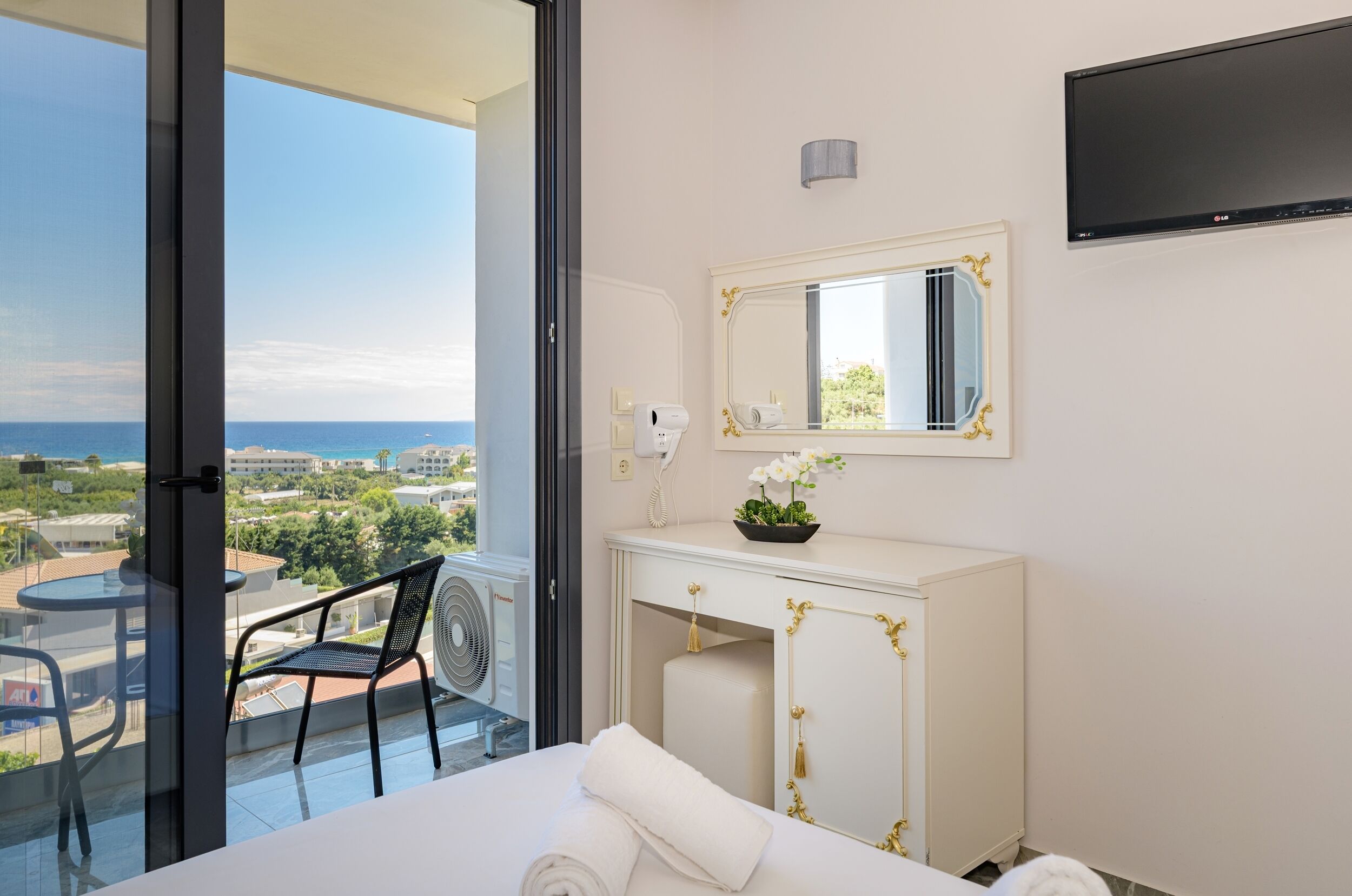 A hotel room with a balcony, a bed, a vanity with a mirror, a TV, and a view of the city and sea.