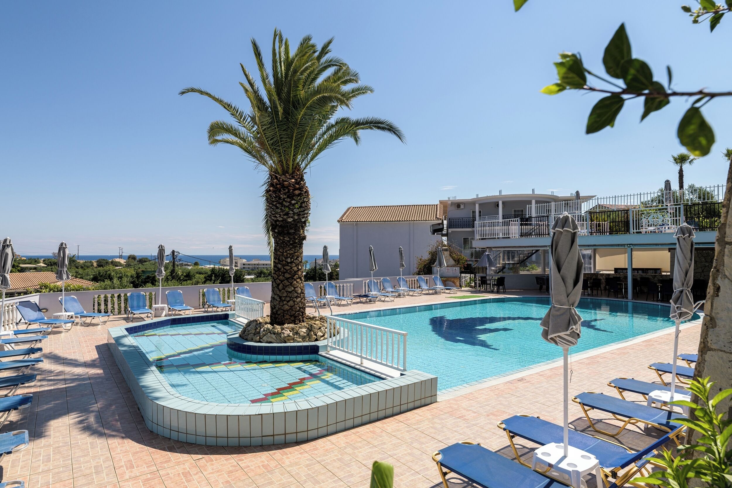 A swimming pool with blue tiles, surrounded by lounge chairs and umbrellas, with a palm tree in the center.
