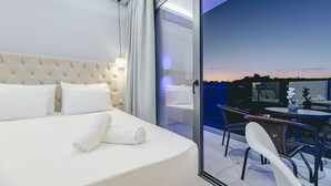 Una habitación de hotel moderna con una cama grande, un ventanal que da a un balcón con mesa y sillas, y vista a la ciudad al atardecer.