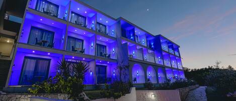 Un hotel de varios pisos con balcones, mobiliario exterior y un camino de piedra que conduce a la entrada.