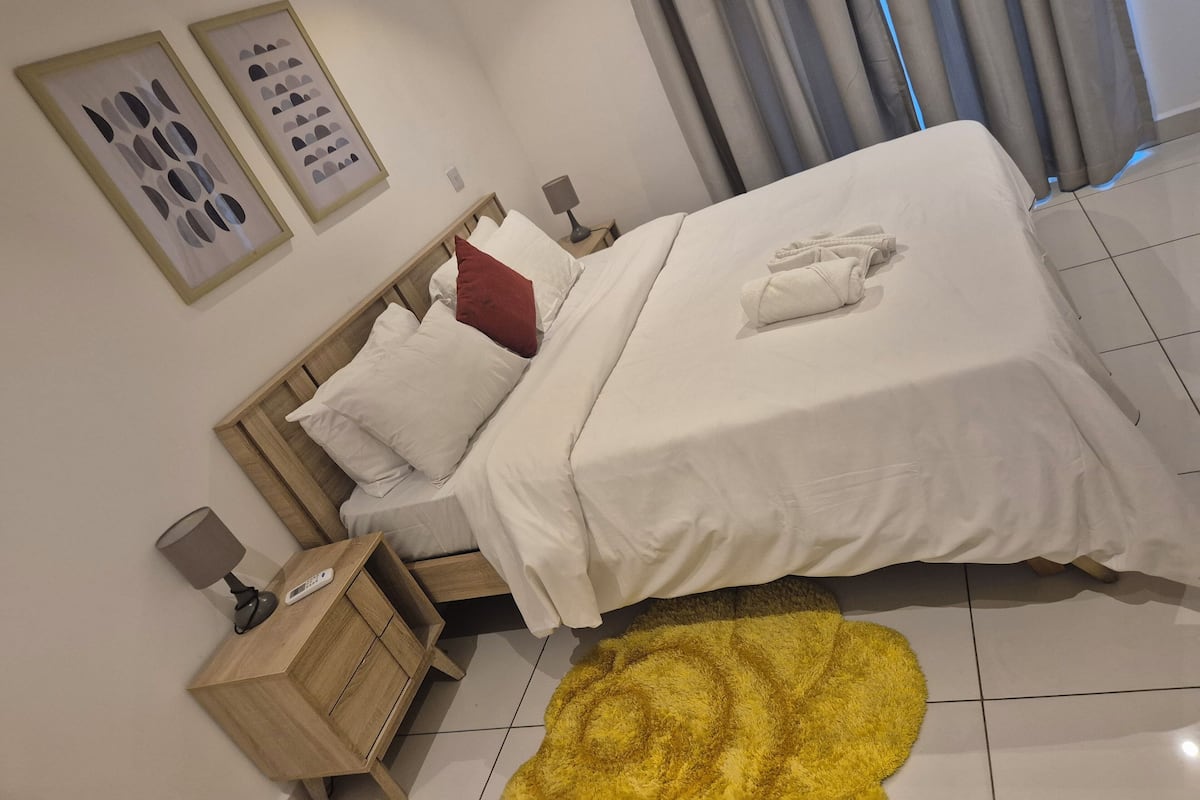 Un lit bien fait, avec du linge de lit blanc et un oreiller rouge, une table de chevet en bois avec une lampe, et un tapis jaune et moelleux sur le sol.