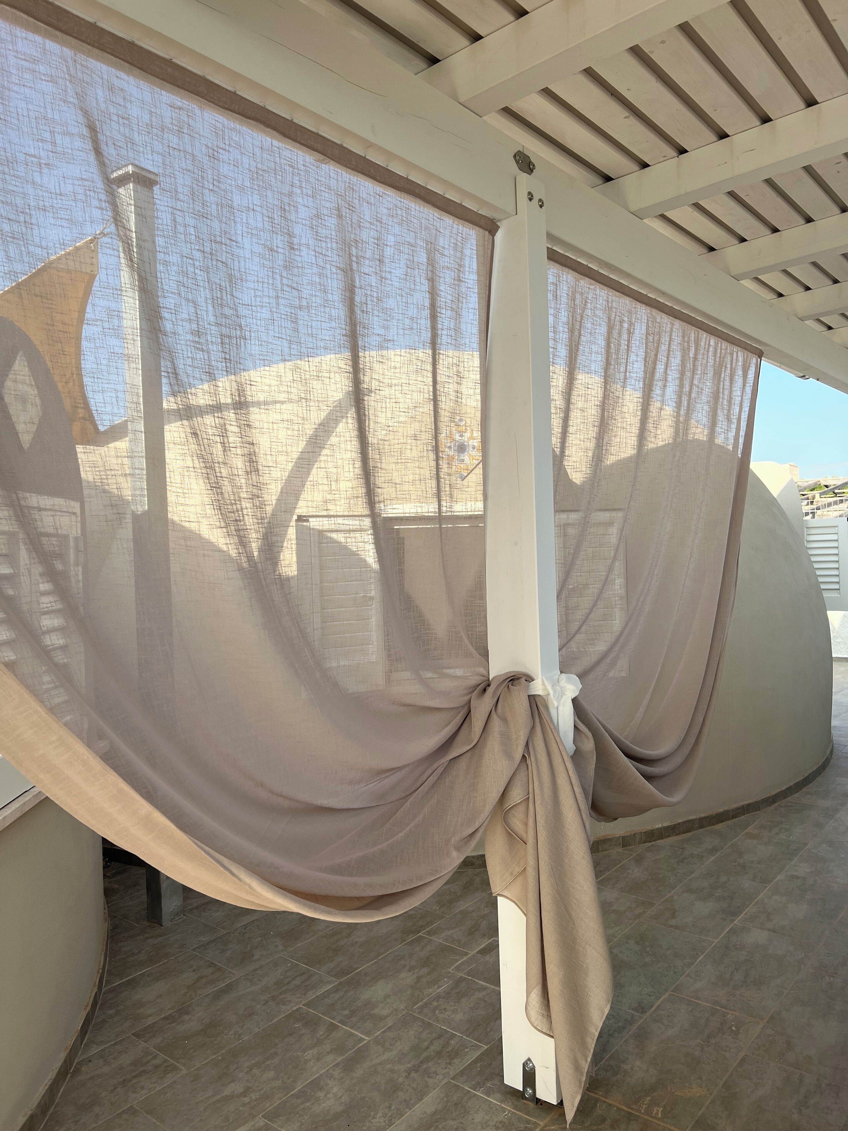 A room with a large window covered by sheer curtains, a white ceiling with beams, and a tiled floor.