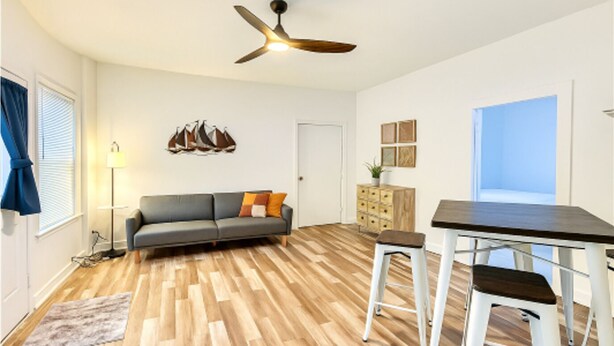 A modern living room with a grey sofa, wooden flooring, a ceiling fan, a dining table with two stools, a lamp, and a decorative wall piece. - The Perfect Getaway 2-bedroom apartment in wonderful Sandusky with WiFi, AC (Sandusky)