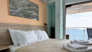 A neatly made bed with white pillows and a beige blanket, a small wooden nightstand, and a view of the sea through an open door. - Agapaki Studios (Neos Marmaras)