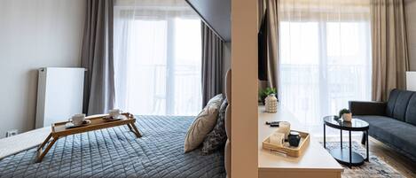 A hotel room with a bed, a bedside table with a tray of breakfast items, a sofa, and a view of the city through large windows.