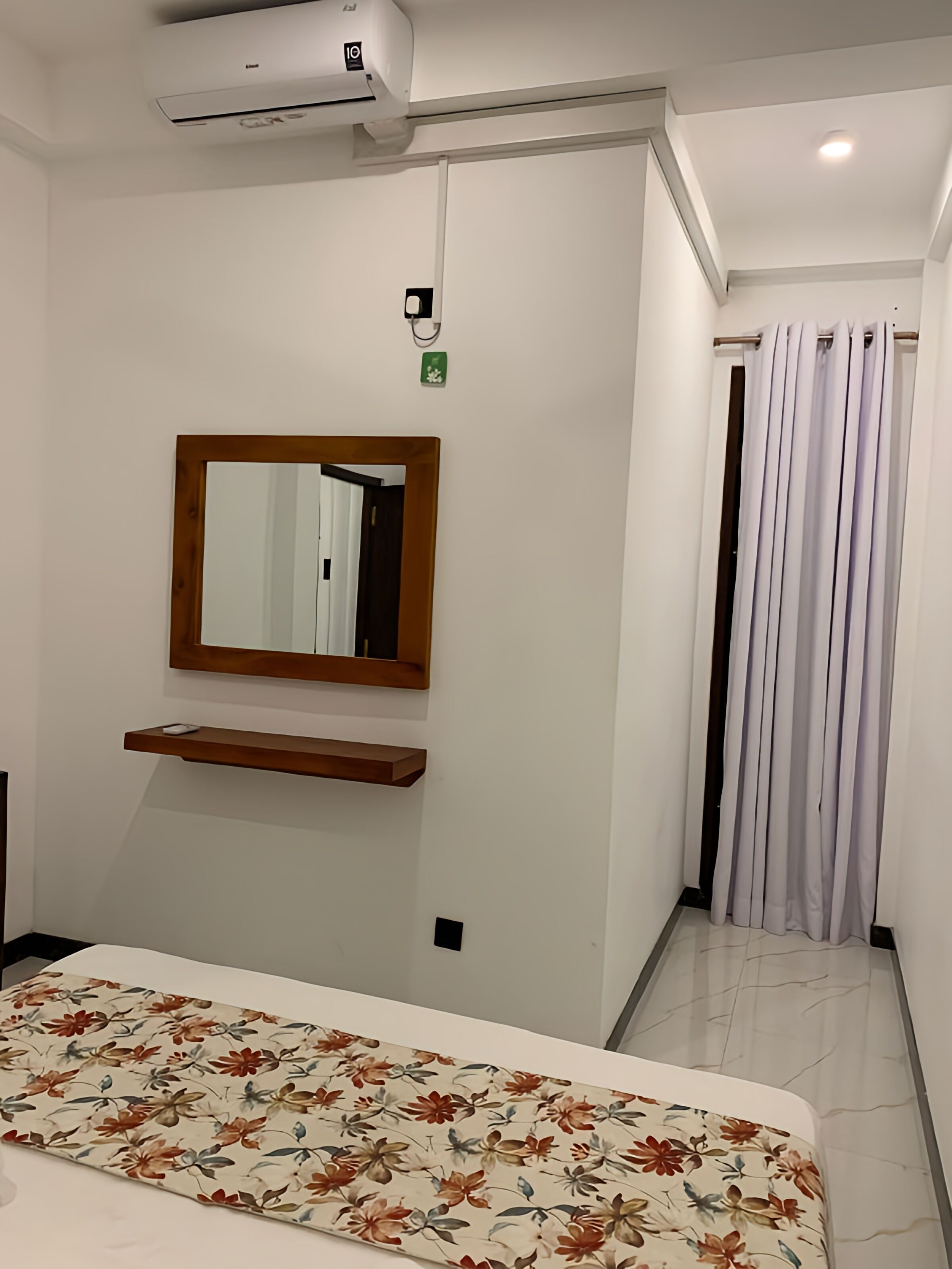 Una habitación de hotel con una cama, un espejo en la pared, un estante, una cortina y un equipo de aire acondicionado.