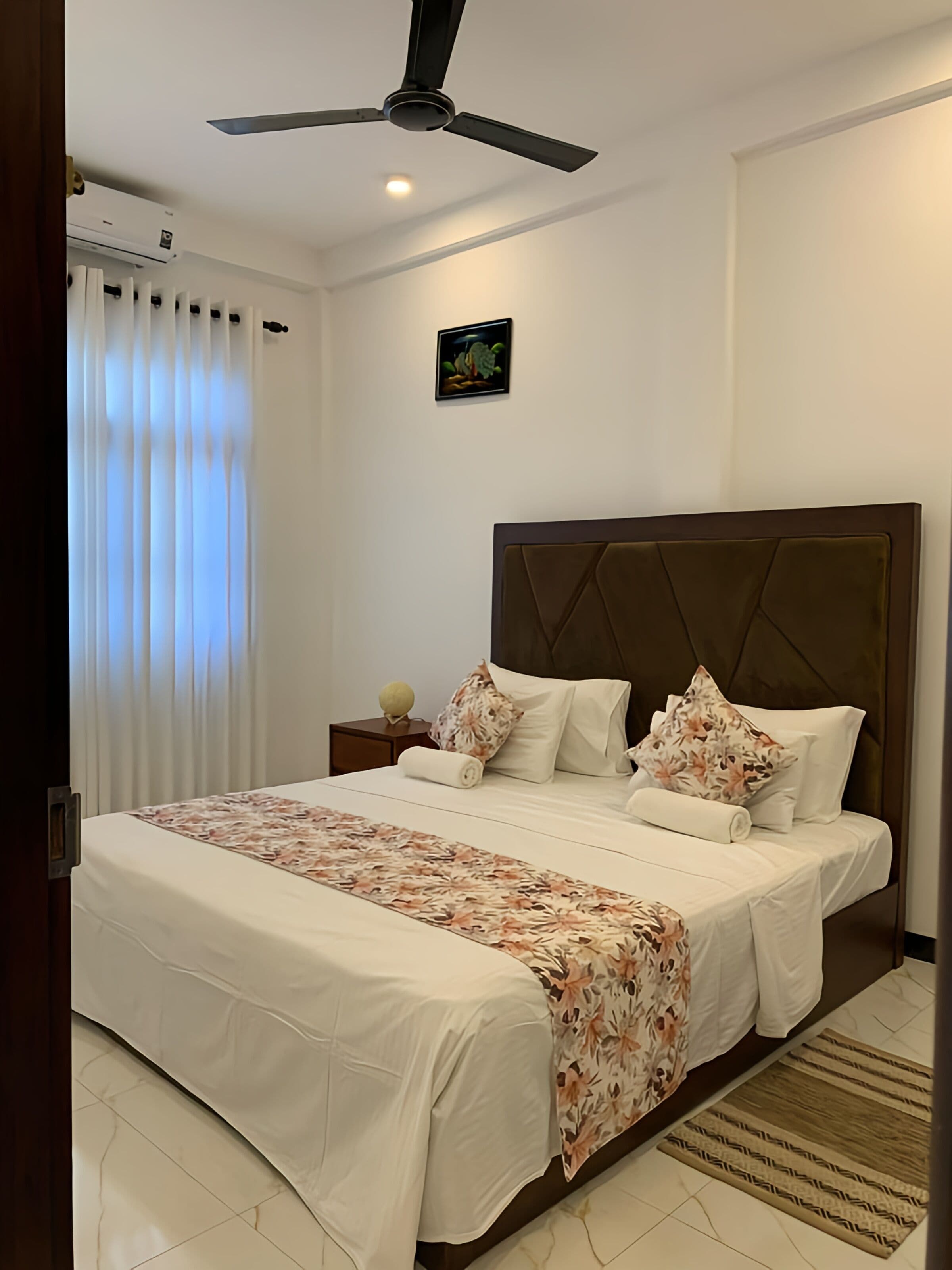 A hotel room with a bed, a ceiling fan, a nightstand, a wall-mounted picture, and a window with curtains.