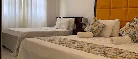 Quarto de hotel com duas camas, cabeceira, mesa de cabeceira, luminária e janela com cortinas.