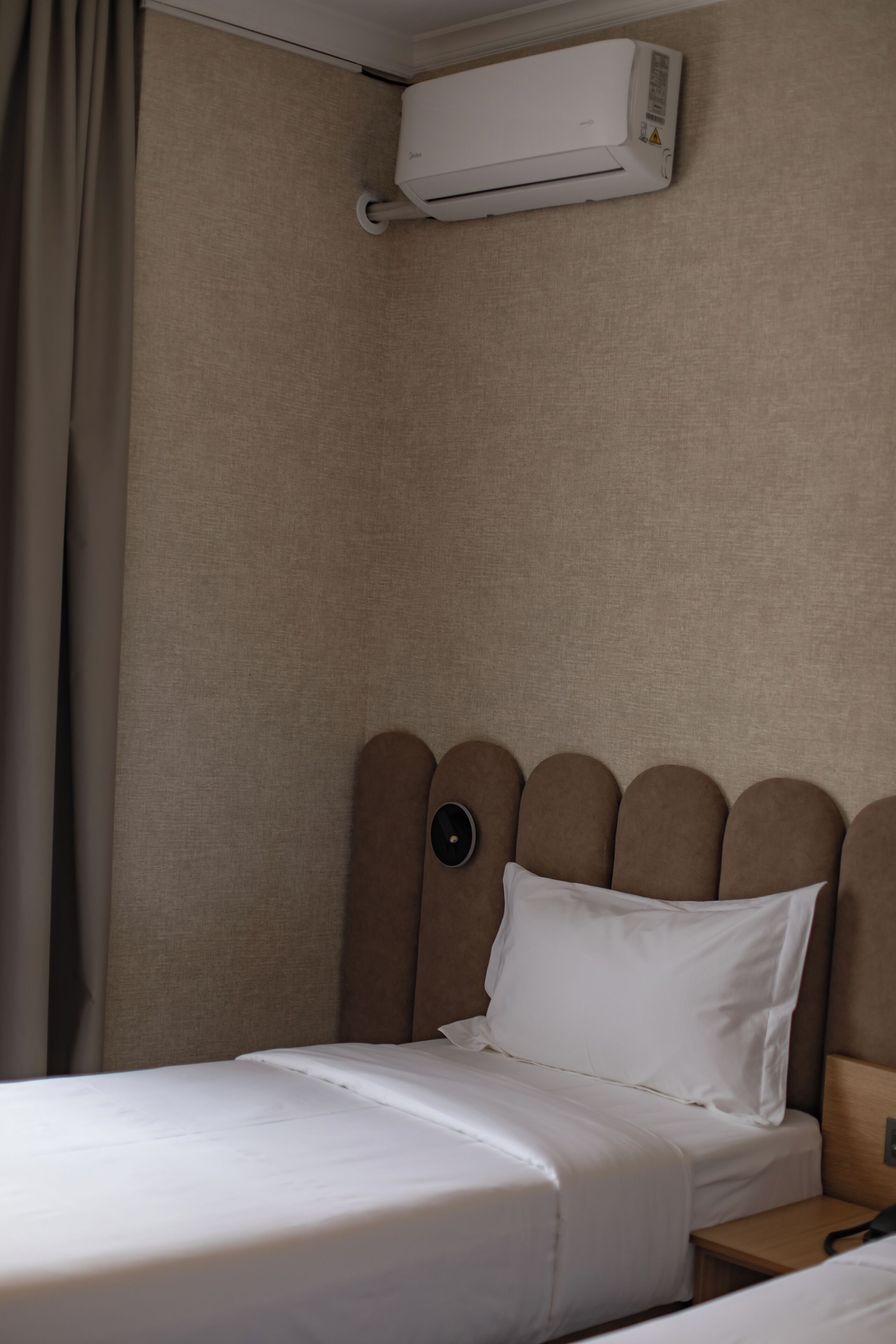 A hotel room with a bed, a nightstand, a wall-mounted air conditioner, and a curtain.