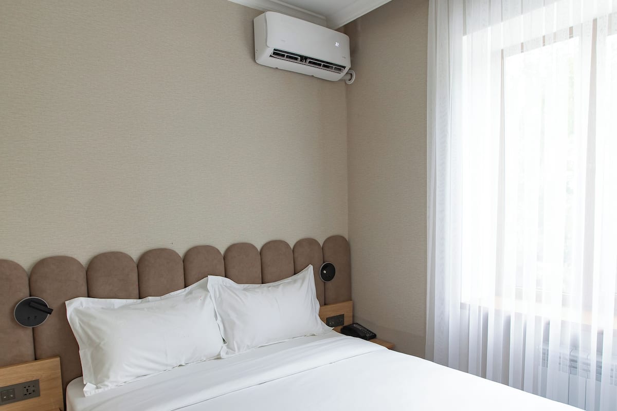 Habitación de hotel con una cama grande, aire acondicionado de pared, una mesita de noche y una ventana con cortinas ligeras.