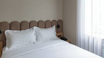 A hotel room with a large bed, an air conditioning unit above the headboard, a bedside table with a lamp, and a window with sheer curtains.