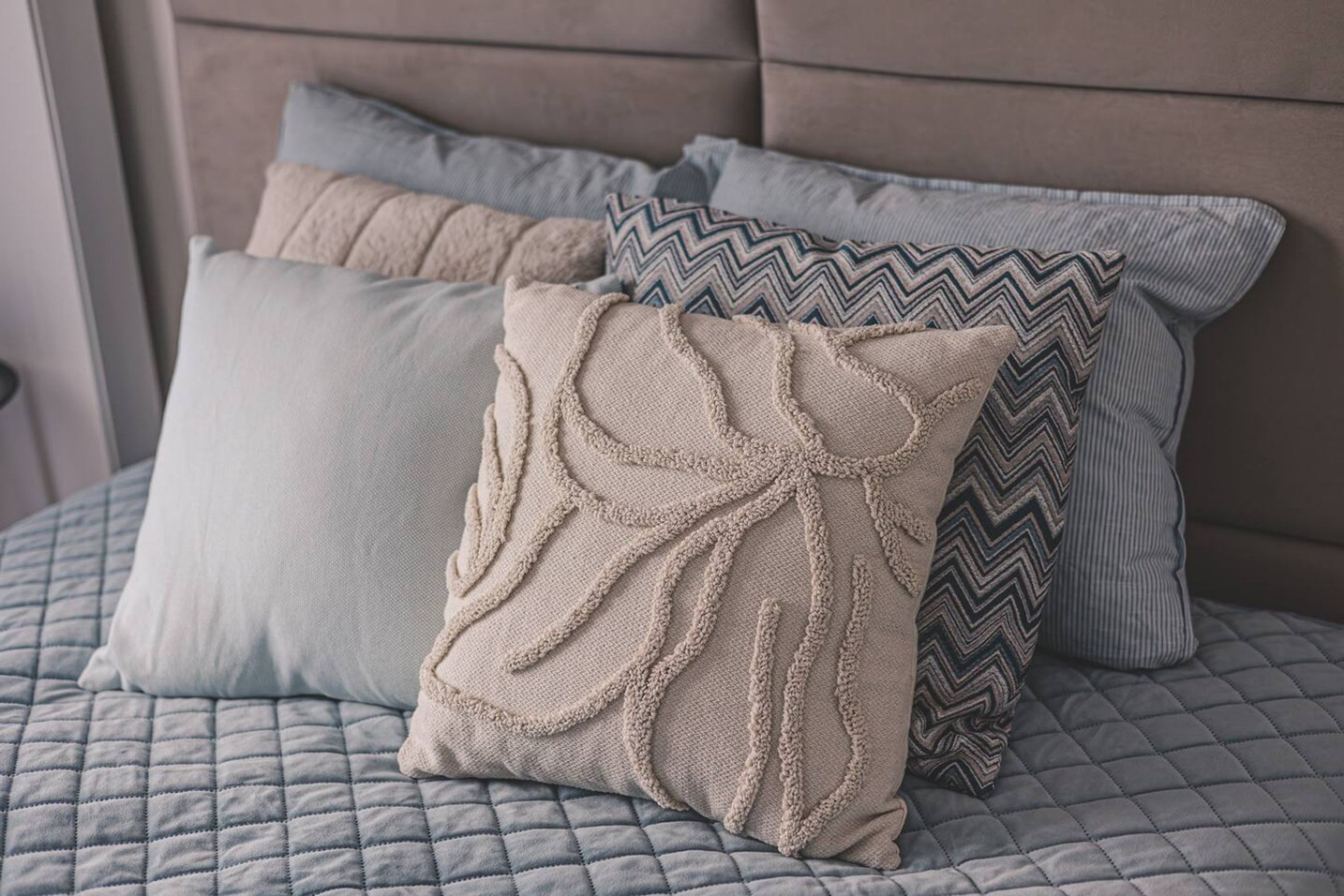 A bed with a quilted grey bedspread and three pillows in various patterns and textures.