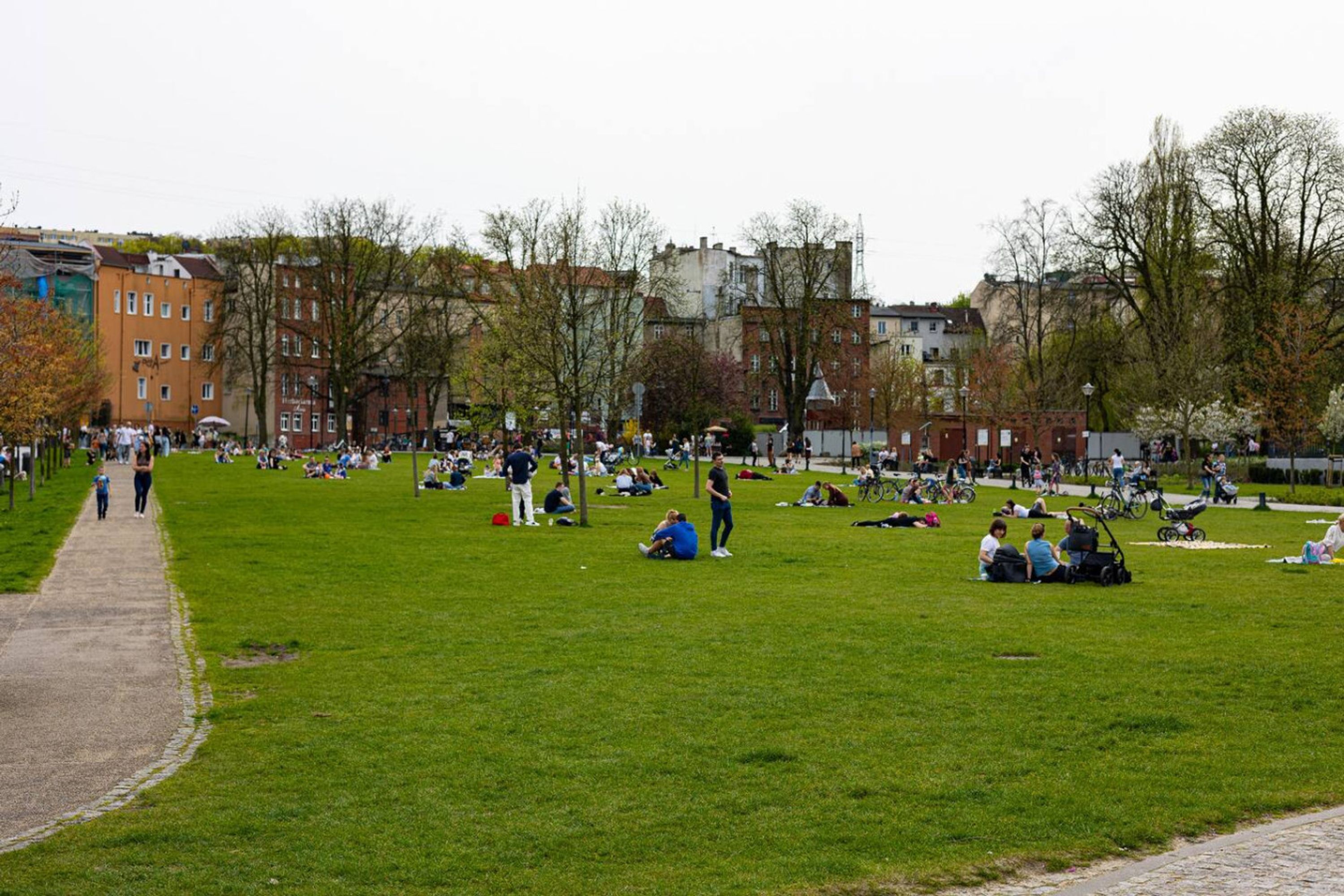 Ein Park mit Menschen, die auf der Wiese sitzen, einem gepflasterten Weg und Gebäuden im Hintergrund.