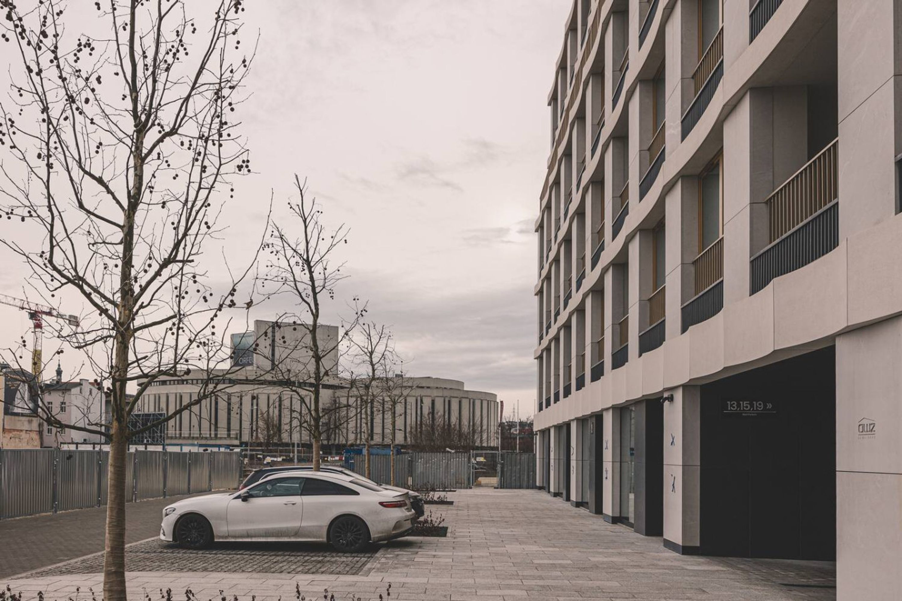 Ein modernes Gebäude mit weißer Fassade, ein Parkplatz mit einem weißen Auto und ein Baum im Vordergrund.