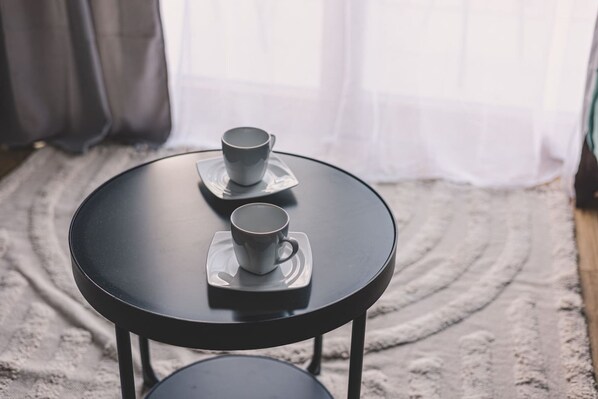 Une table ronde noire sur laquelle reposent deux tasses blanches sur des soucoupes, un tapis à motifs et, en arrière-plan, un rideau blanc et léger.