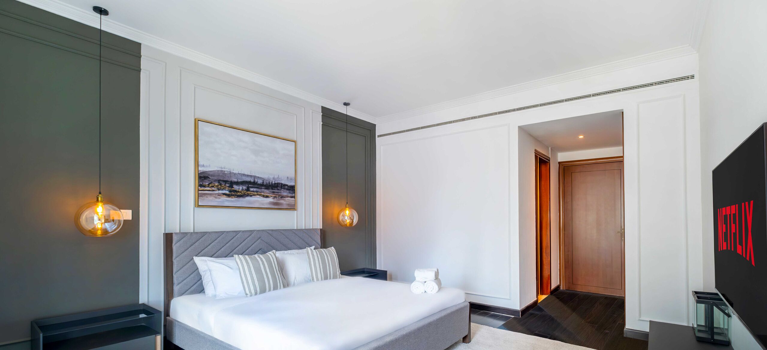 Quarto de hotel moderno com uma cama grande, abajures, uma moldura com um quadro na parede e uma porta que dá para outro cômodo.