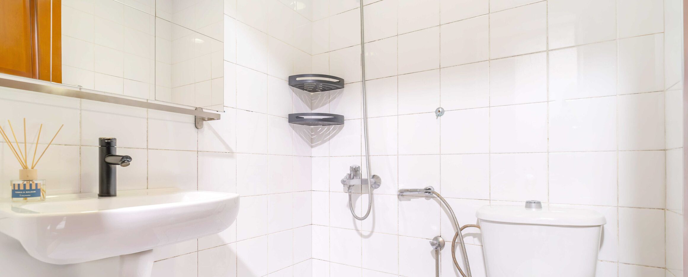 Kylpyhuoneessa on valkoiset laatat, valkoinen pesuallas, wc-istuin ja suihku, jossa on käsisuihkupää.