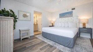 Un dormitorio con cama, mesitas de noche, una silla de mimbre, un cuadro enmarcado y un acceso a baño.