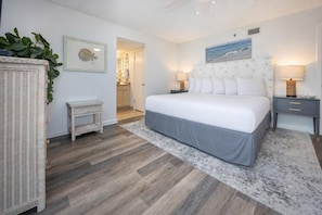A bedroom with a bed, bedside tables, a nightstand, a wicker chair, a framed picture, and a doorway leading to a bathroom. - Phoenix East 2 2065 3 Bedroom Condo (Orange Beach)