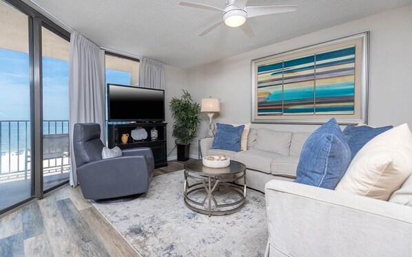 A modern living room with a flat-screen TV, a gray sofa, a round coffee table, and a large window overlooking a beach. - Phoenix East 2 2065 3 Bedroom Condo (Orange Beach)
