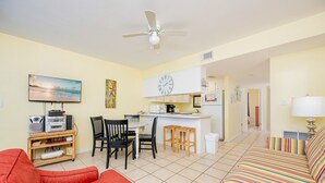 A living room with a red sofa, a TV, and a ceiling fan. There is a kitchen area with a clock and a refrigerator. - Island Winds West 876 1 Bedroom Condo (Gulf Shores)