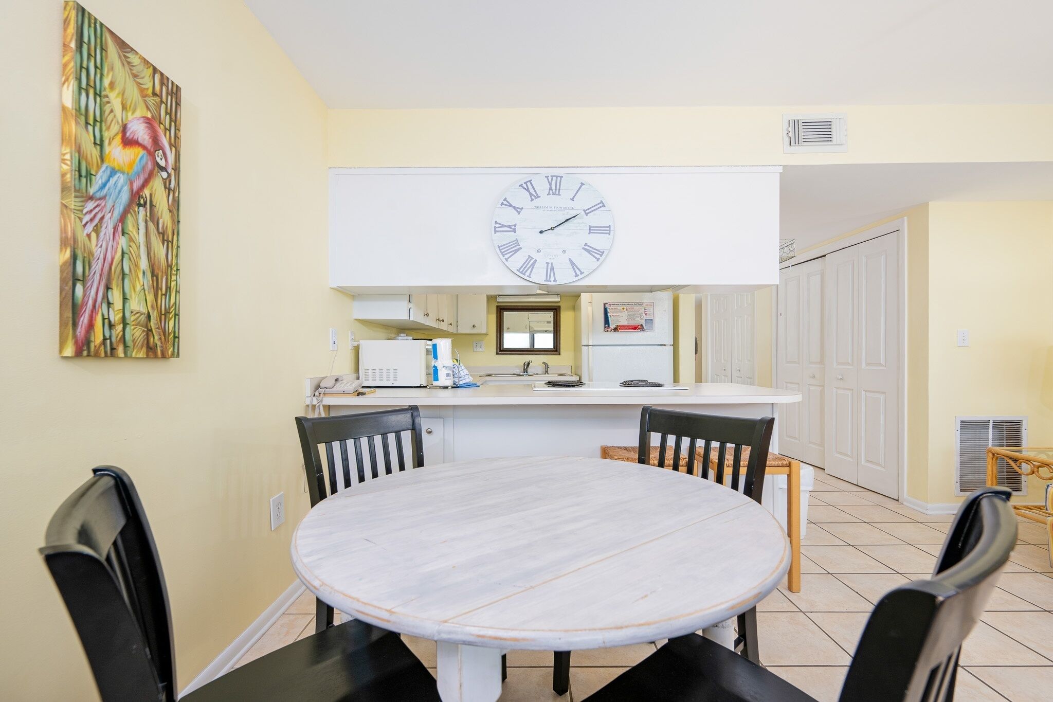 A dining area with a round table and four chairs, a clock on the wall, and a colorful painting of a parrot.