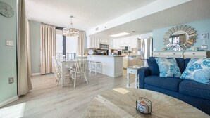 A modern living room with a blue sofa, a dining area with a table and chairs, a kitchen with white cabinets, and a large mirror on the wall.