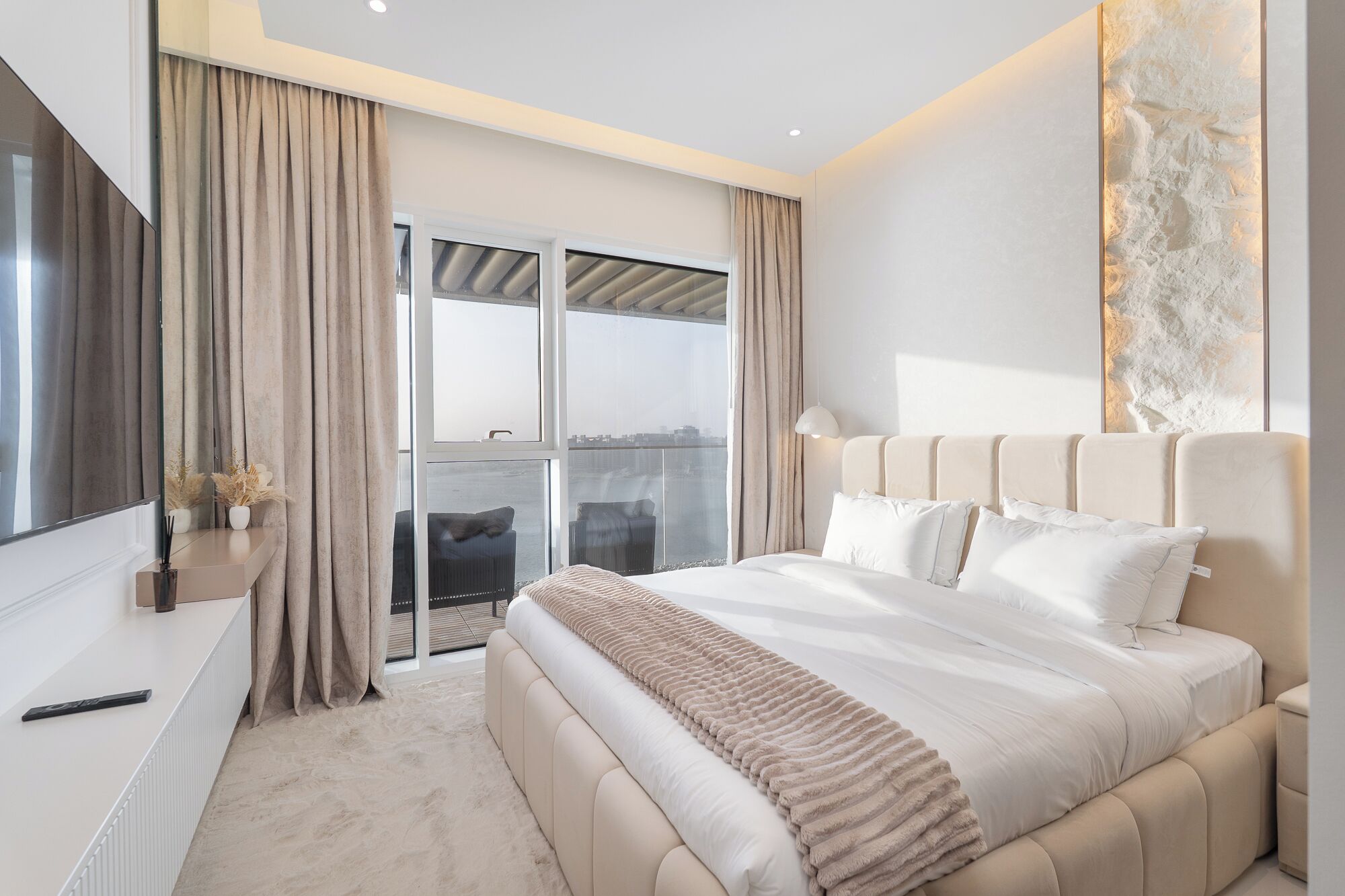 A modern bedroom with a large bed, a view of the city through floor-to-ceiling windows, and a balcony.