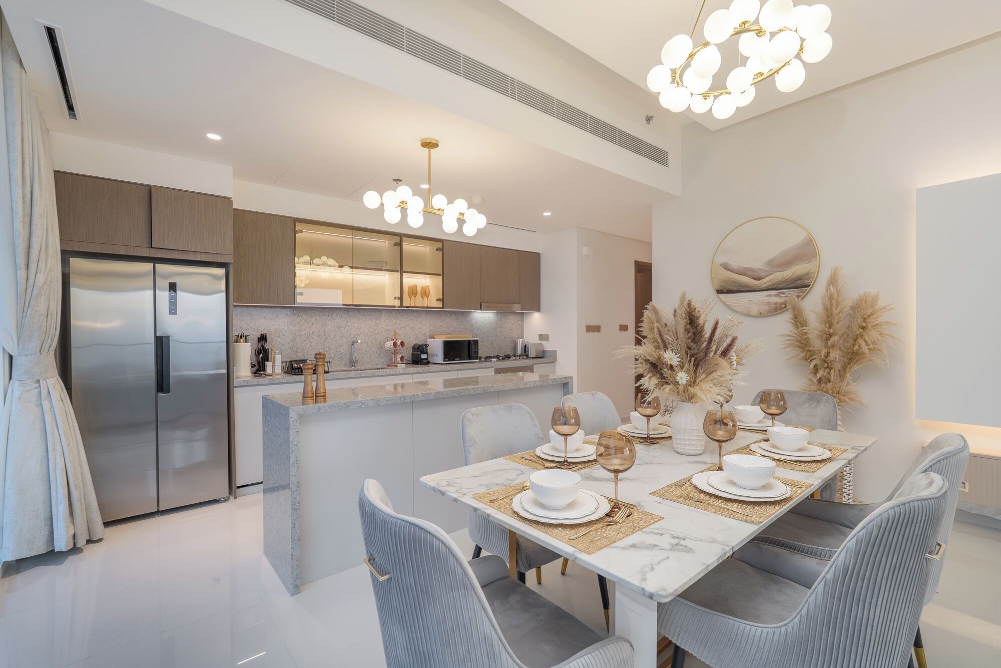 A modern kitchen with a built-in island, stainless steel appliances, and a dining area set for six with a marble table and grey upholstered chairs.