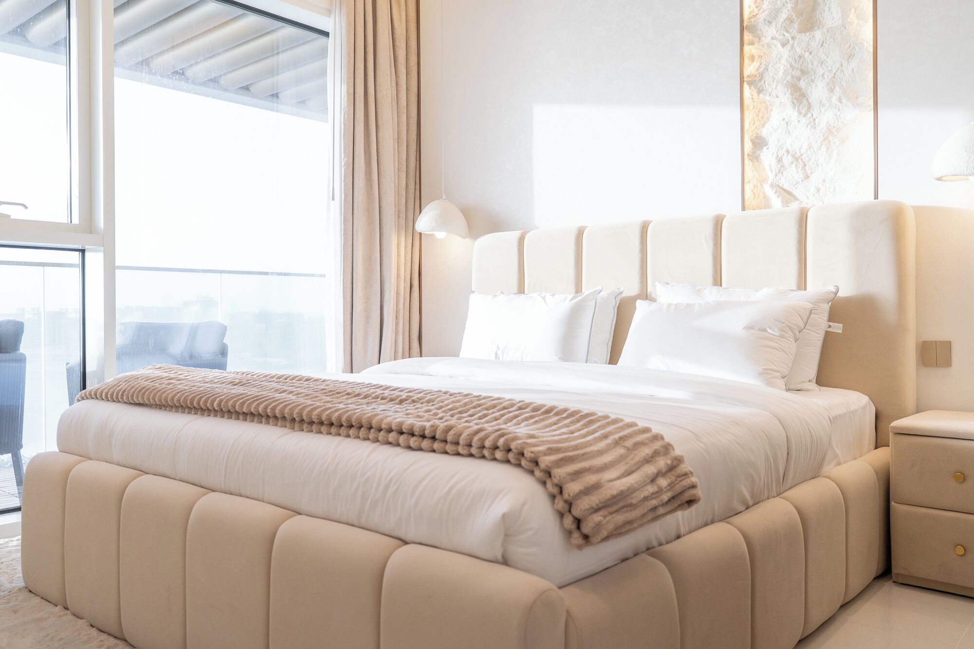 A modern bedroom with a large bed, a nightstand, and a view of the city through a large window.