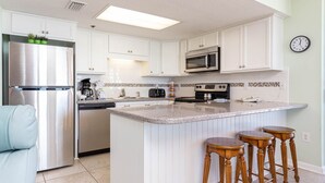 A modern kitchen with white cabinetry, stainless steel appliances, a large island with bar stools, and a clock on the wall. - Phoenix East 2 2105 3 Bedroom Condo (Orange Beach)