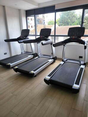 Three treadmills in a room with large windows, wooden flooring, and a view of buildings and trees outside. - leo residence (Nairobi)