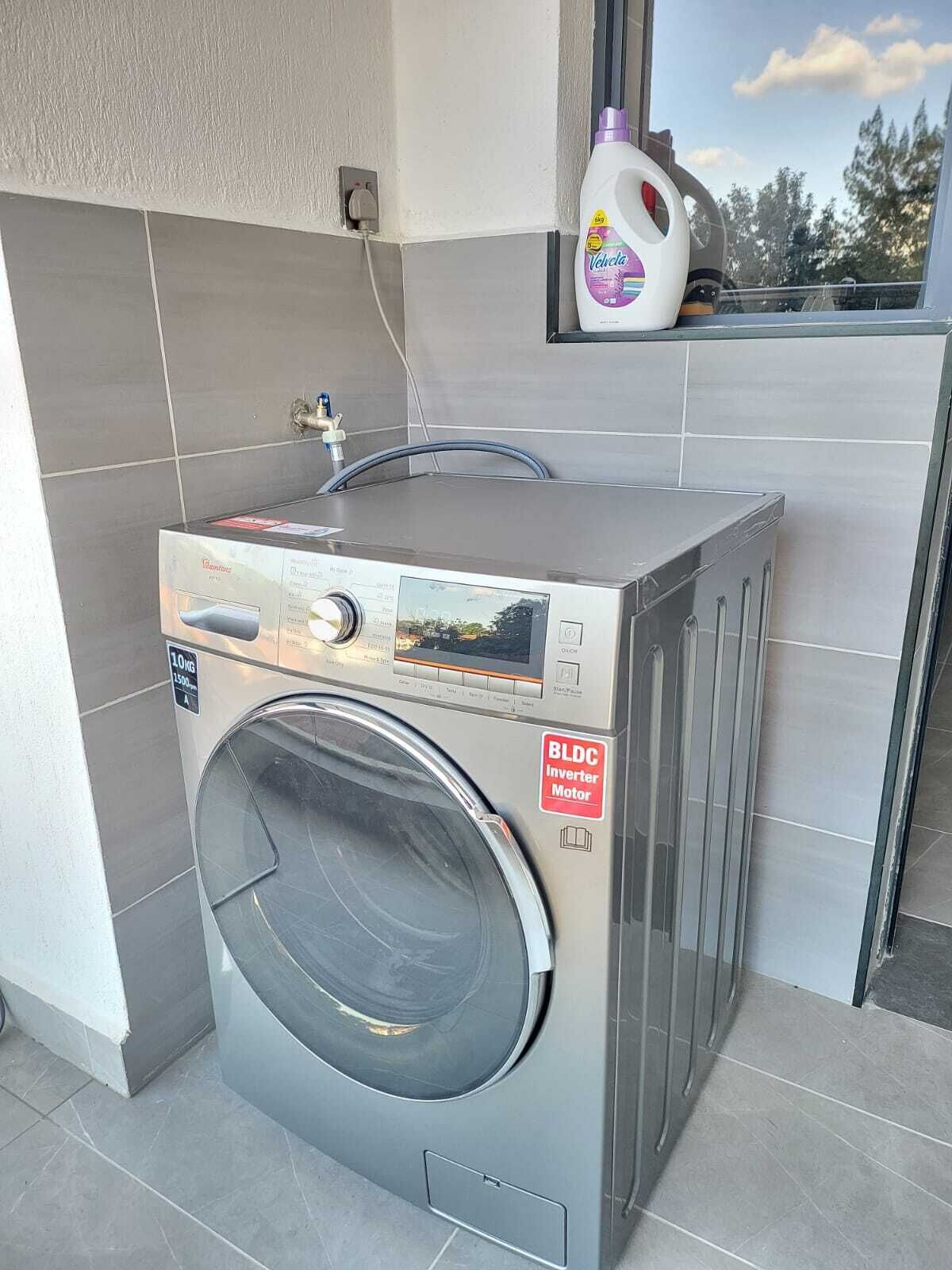 A compact, modern front-loading washing machine with a BLDC inverter motor, placed in a clean, tiled laundry area with a window above it.