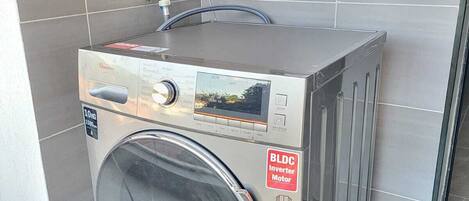 A compact, modern front-loading washing machine with a BLDC inverter motor, placed in a clean, tiled laundry area with a window above it.