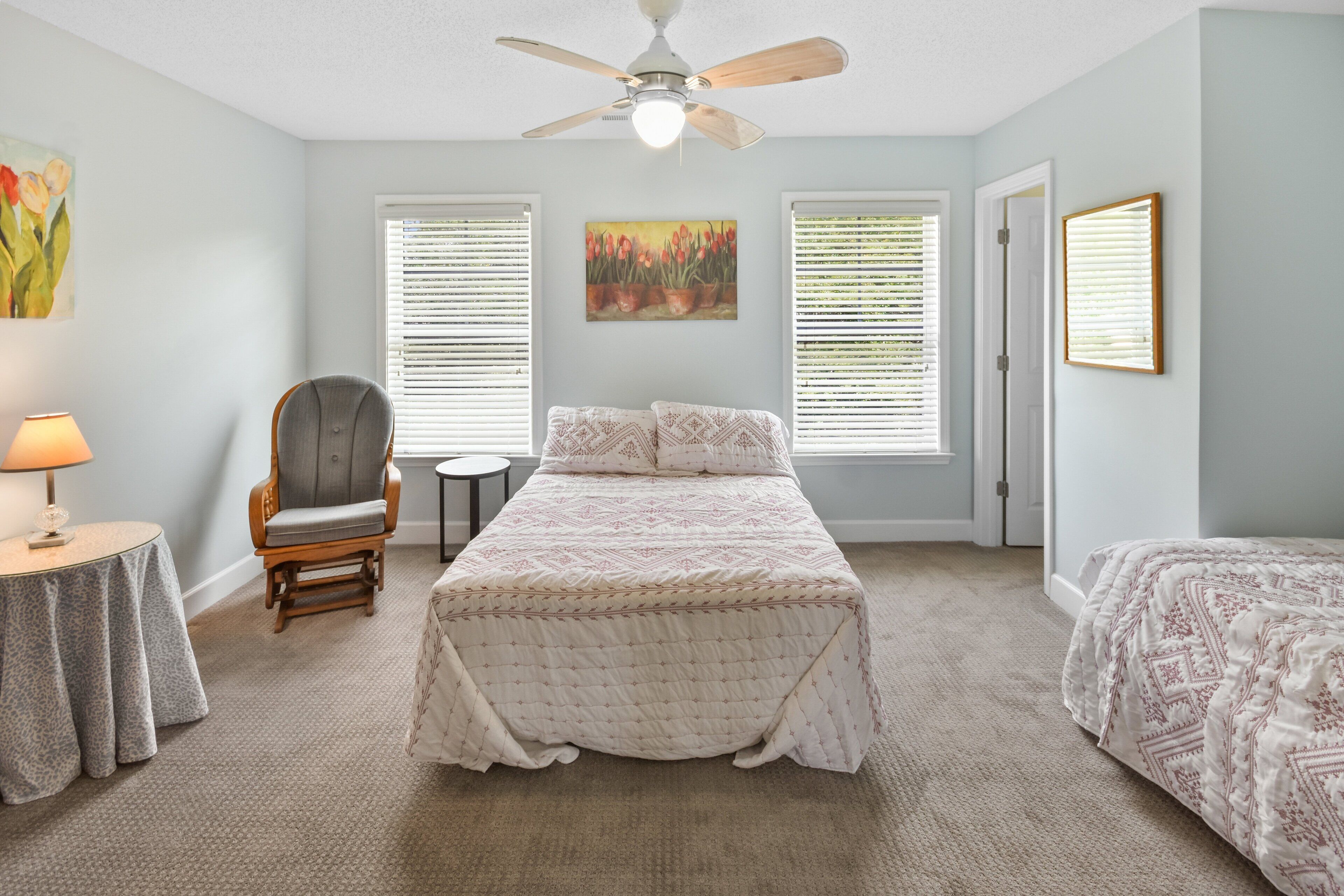 A bedroom with a bed, a chair, a table, and two windows with blinds.